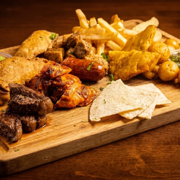 Mixed grilled meat platter with flatbreads, fries, pastries, potatoes, vegetables, lime, and dipping sauces on a wooden serving board.
