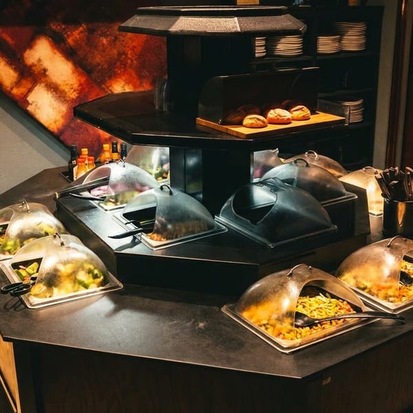 A dark, hexagonal buffet table with covered food trays, bread rolls, and stacked plates in a restaurant.