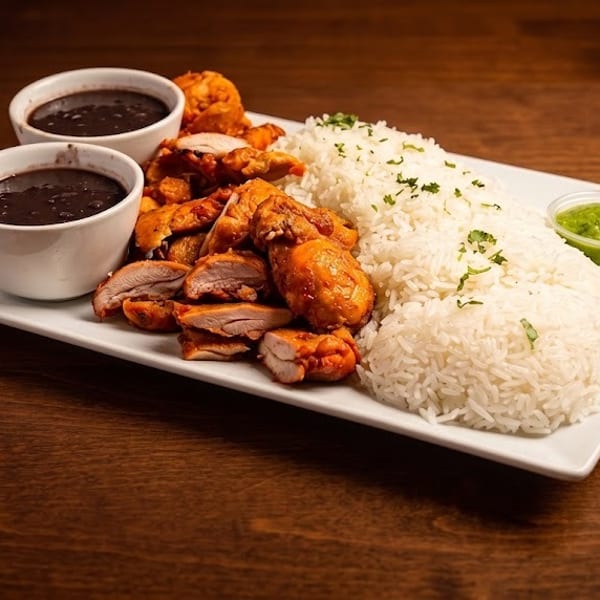Plate of grilled chicken, white rice, black beans, and green sauce on a wooden table.