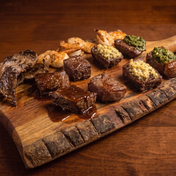 Assortment of grilled meats, shrimp, and sauced steak bites served on a rustic wooden board.