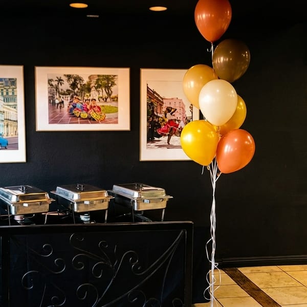 Buffet setup with chafing dishes, Cuban-themed art, and balloons in warm colors.