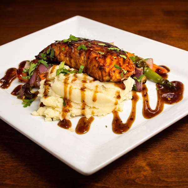 Glazed salmon fillet with mashed potatoes, sautéed vegetables, brown sauce, and fresh herbs on a white plate.