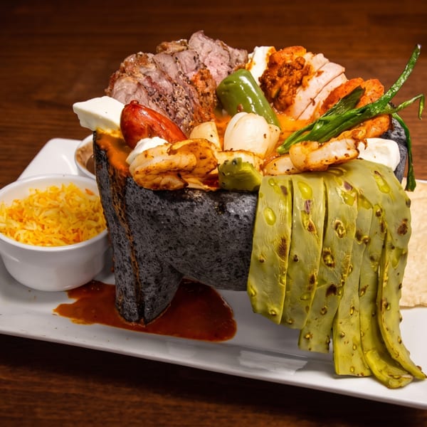 Mixed grilled meats, shrimp, vegetables, pickles, rice, tortillas, and sauce served on a white platter.